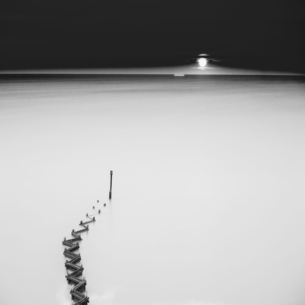 Black and white photograph of an uneven line of wooden posts curving through smooth pale water, with a distant bright light beneath a dark sky.