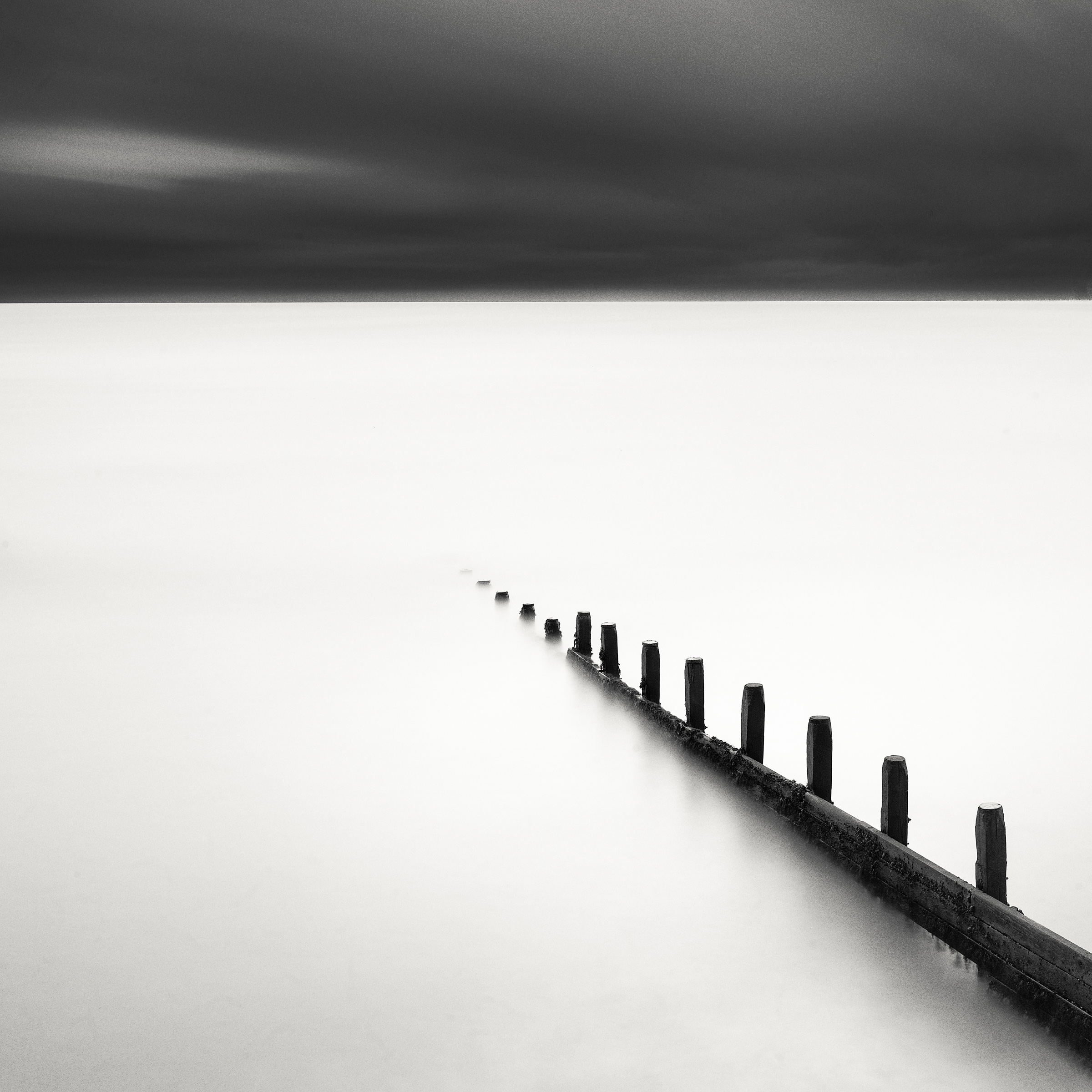 Black and white photograph of a wooden groyne running diagonally from the lower right into smooth pale water beneath a dark sky.