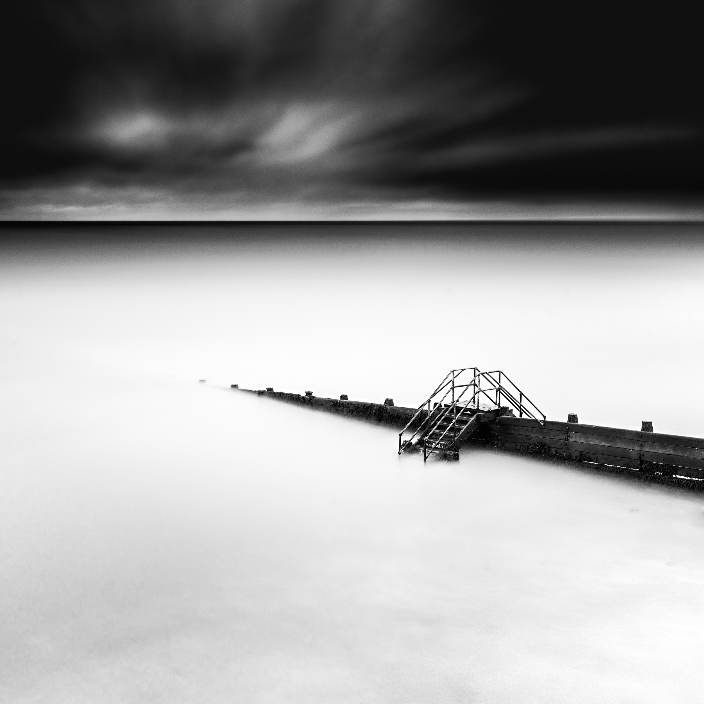 Black and white photograph of metal steps crossing a coastal groyne, with smooth pale water and a dark, clouded sky.