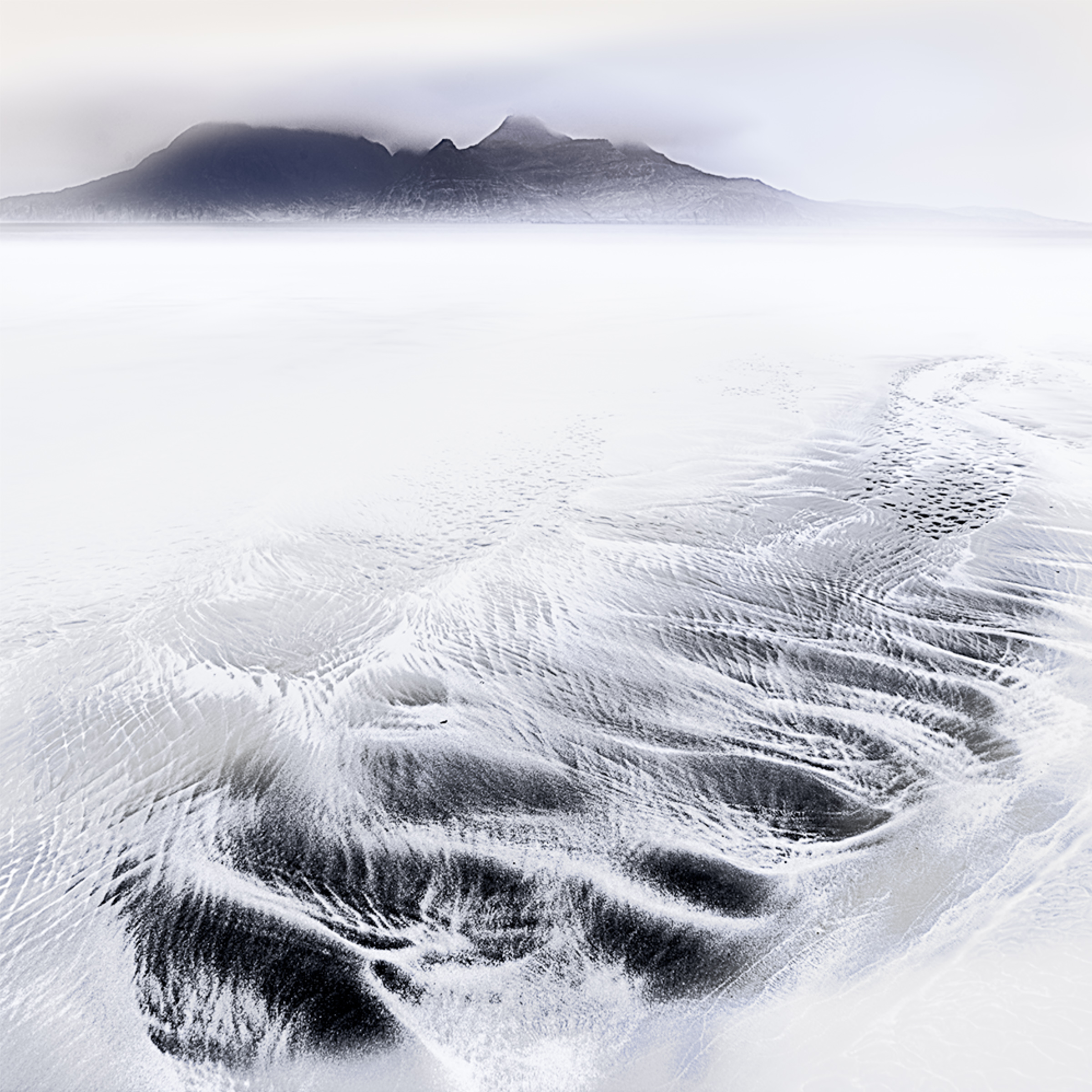 Rippled sand and shallow water at Laig Bay, Isle of Eigg, with distant mountains partly veiled by cloud.