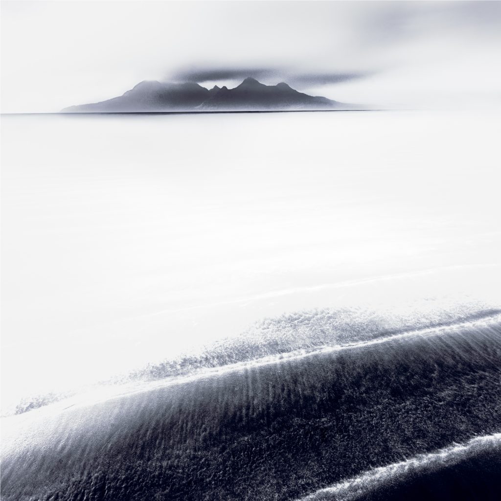 Dark diagonal bands of sand beneath pale water, with a cloud-covered mountain on the horizon.
