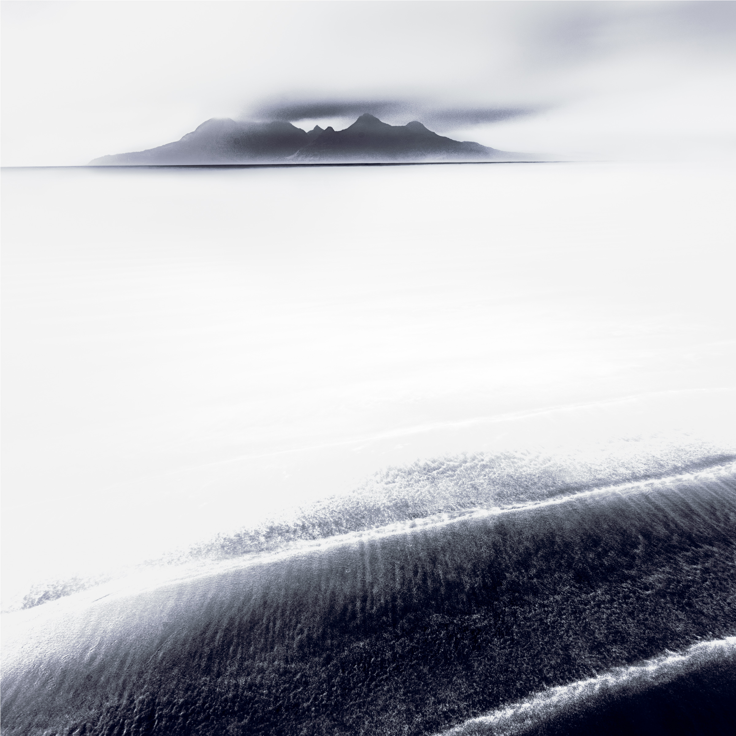 Dark diagonal bands of sand beneath pale water, with a cloud-covered mountain on the horizon.
