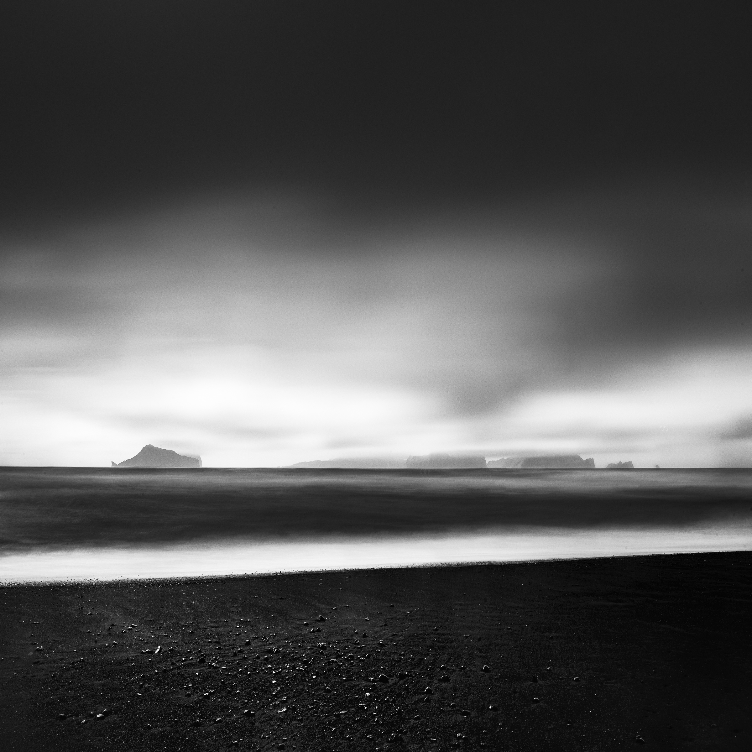 Black and white view towards the Westman Islands beneath an expansive dark sky, with blurred surf crossing a black sand beach.