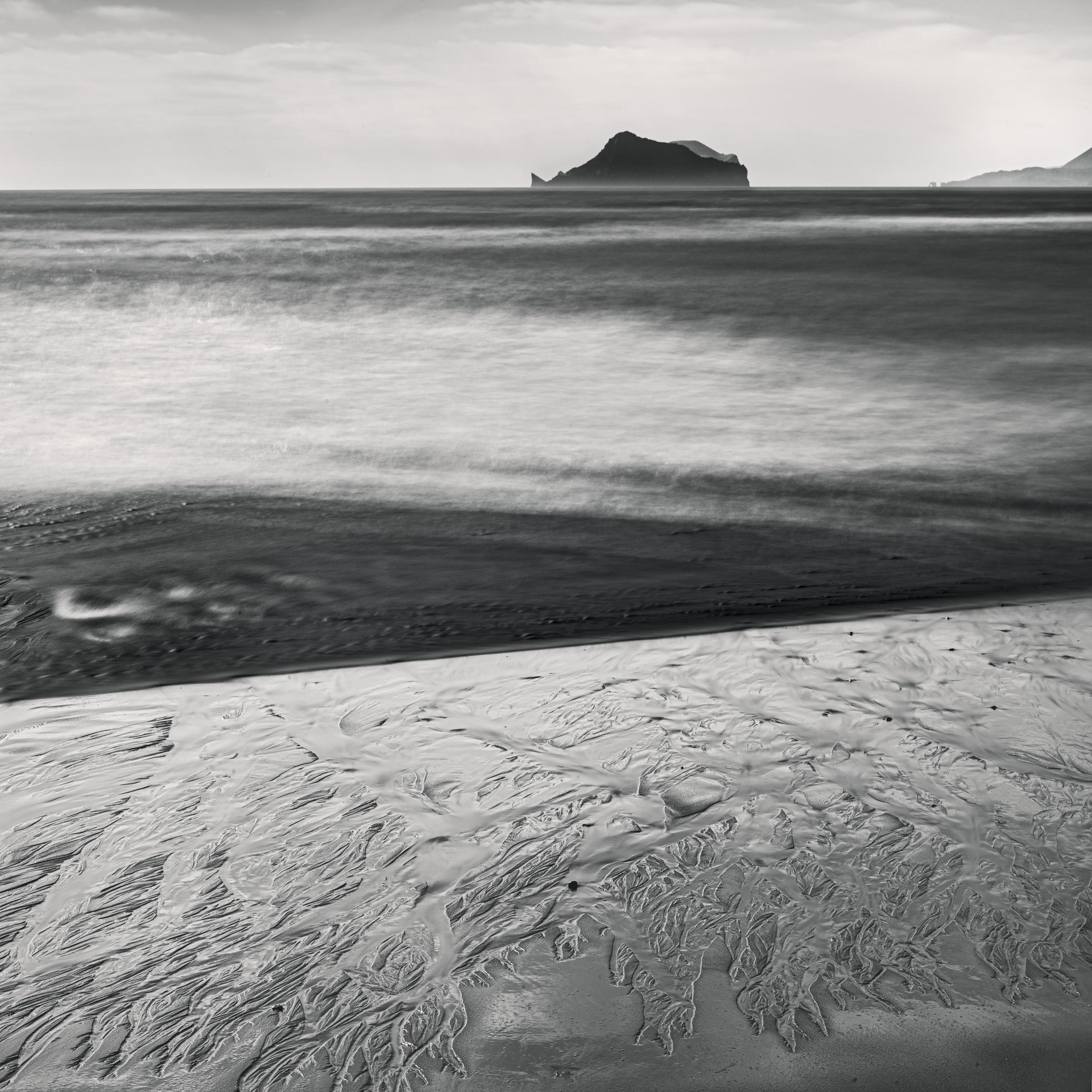 Black and white photograph of branching patterns in wet sand, with rolling surf and a distant island off Iceland’s south coast.