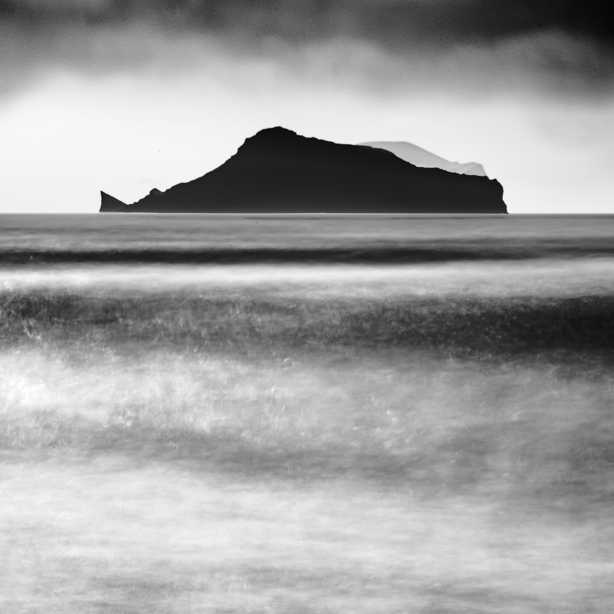 Black and white photograph of a dark island silhouette beyond softly blurred waves, viewed from Iceland’s south coast.
