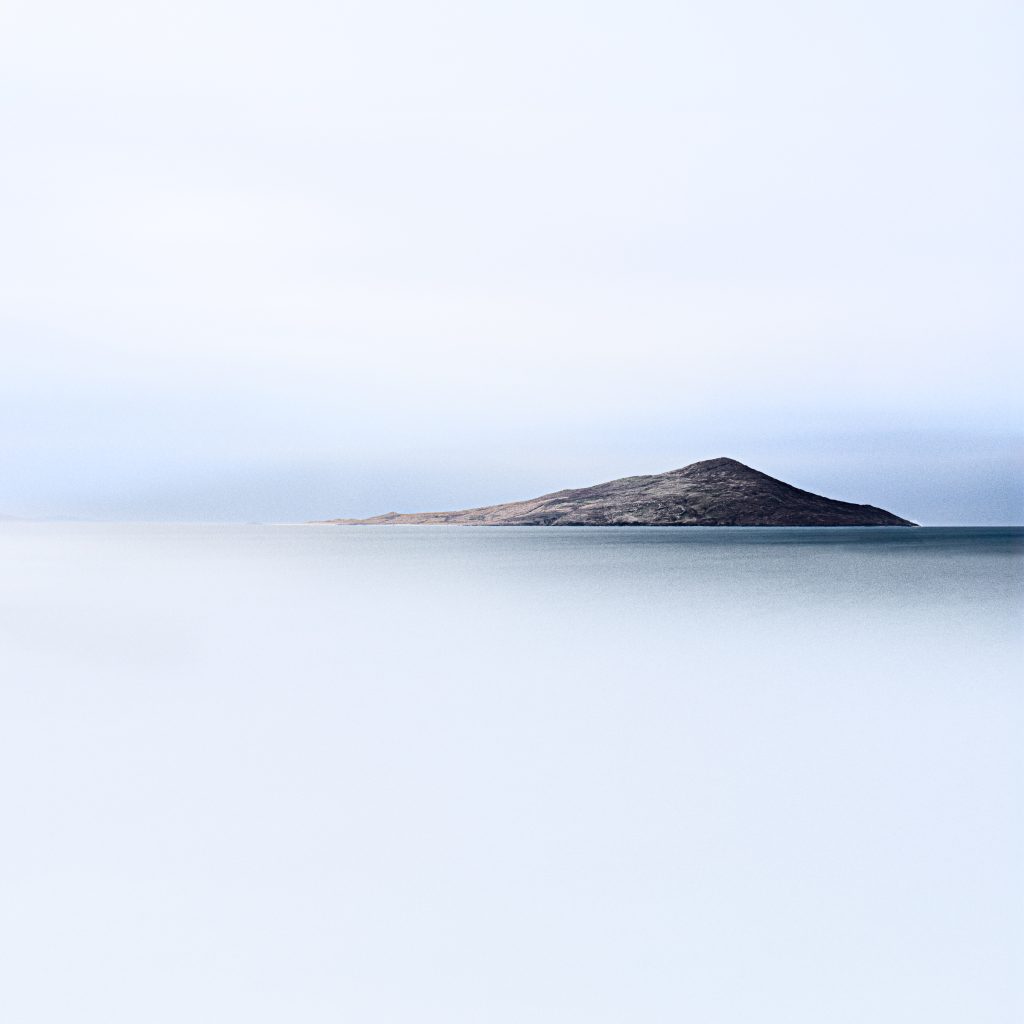 A low rocky island to the right of the frame, surrounded by smooth pale blue water beneath a light grey sky.