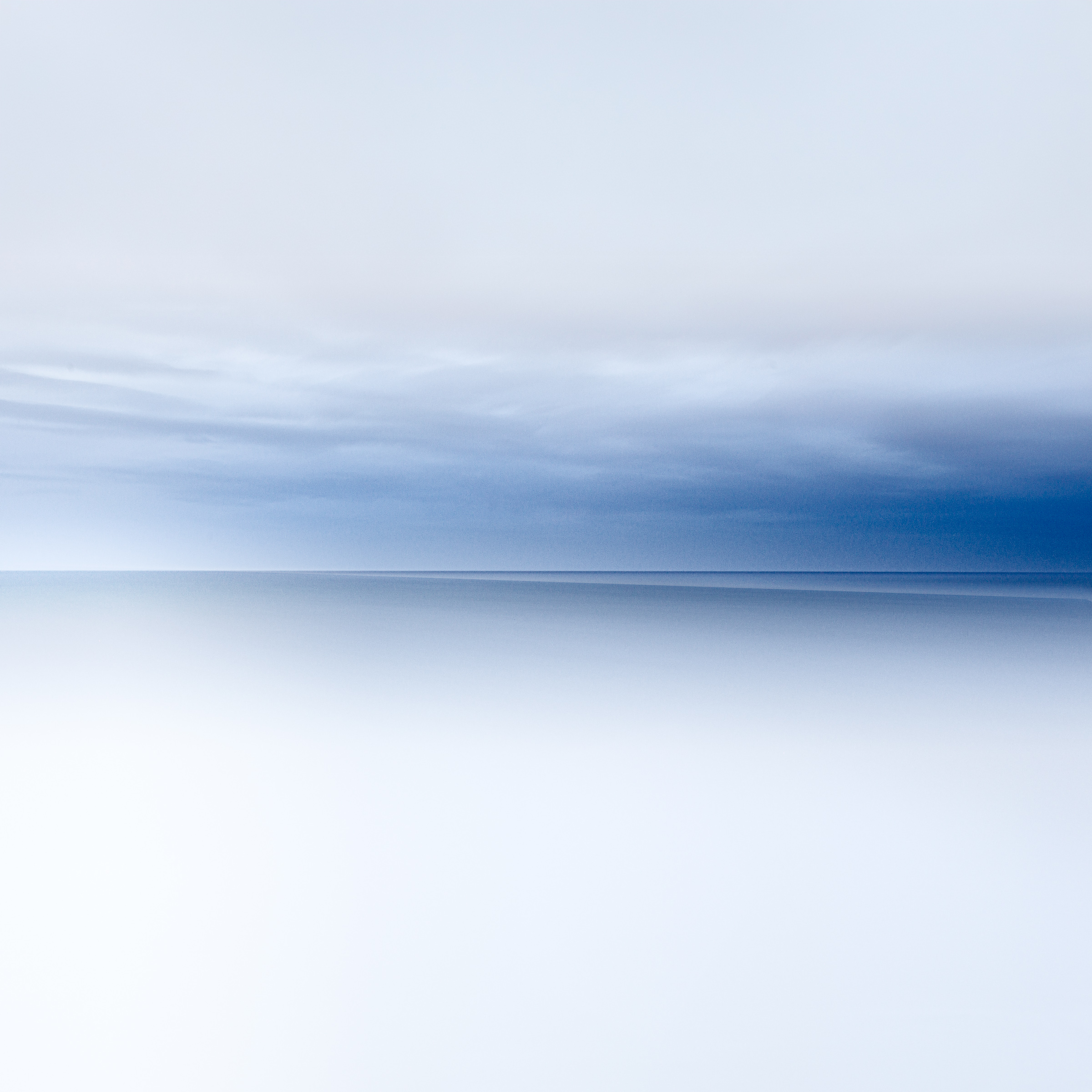 Soft blue and grey clouds above a level sea horizon, with pale, smooth water in the foreground.
